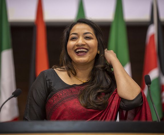 A woman in traditional dress stands in front of various flags.